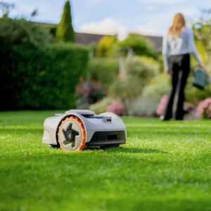 the Segway Navimow i105E Robotic Mower mowing a lawn with a woman watering flowerbeds in the background