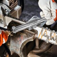 someone sharpening a chainsaw held in a vice, using a chainsaw file and file guide