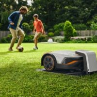 a STIHL iMow robotic lawn mower on its charging station, with a father and son playing football in the background