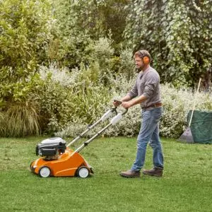 a man wearing ear defenders, scarifying a lawn with an orange STIHL scarifier