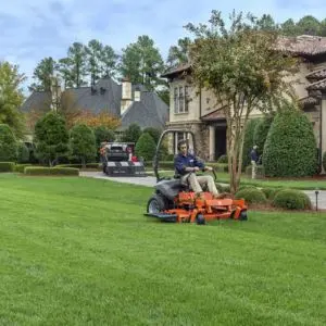 a man mowing the lawn outside a large house, using an orange Husqvarna zero turn lawnmower
