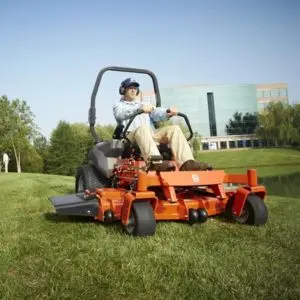 a man cutting a playing field using a zero turn mower, with trees and a high-rise building in the background