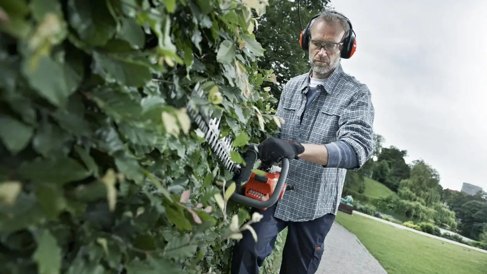 a man wearing ear defenders, safety glasses and gloves, using a hedge trimmer