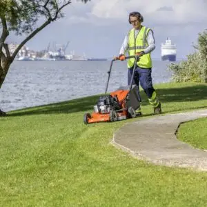 a woman in a yellow, high-viz jacket, mowing grass near the coast, with a ship and dockyard in the background