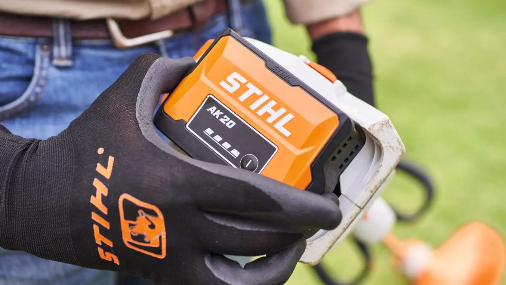 someone wearing a black and orange STIHL-branded glove, holding a STIHL AK 20 battery