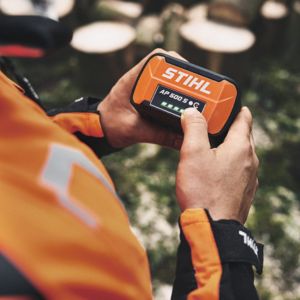a close up over the shoulder of a man dressed in an orange and black jacket, holding a STIHL AP 500 S battery