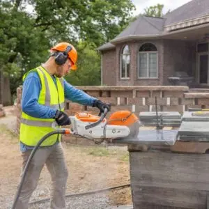 a man wearing an orange hardhat, ear defenders, high-visibility jacket, and gloves, operating the STIHL TSA 300 Cordless Cut-off Machine outside a house