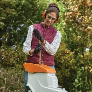 a woman wearing ear defenders, safety glasses and gloves, feeding a small branch into a chipper shredder