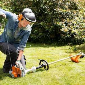 a man wearing visor, ear defenders and gloves, pulling the starting cord of a petrol strimmer that is laid on a lawn