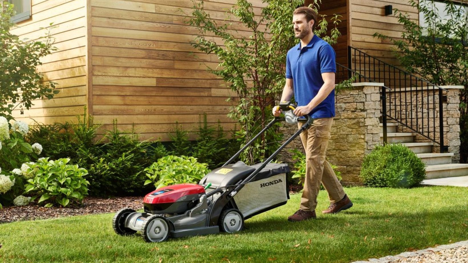 a man dressed in a blue polo shirt and beige trousers, mowing a lawn with a red Honda lawnmower