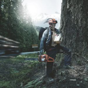 a man stood next to a tree, holding a Husqvarna 262 XP® Chainsaw, looking over his shoulder at passing traffic on a road