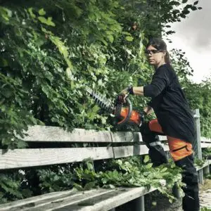 a woman leaning against a bench and using a Husqvarna hedge cutter to trim the side of a hedge