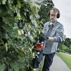 a man wearing safety glasses and ear defenders, cutting the side of a hedge with a hedge trimmer