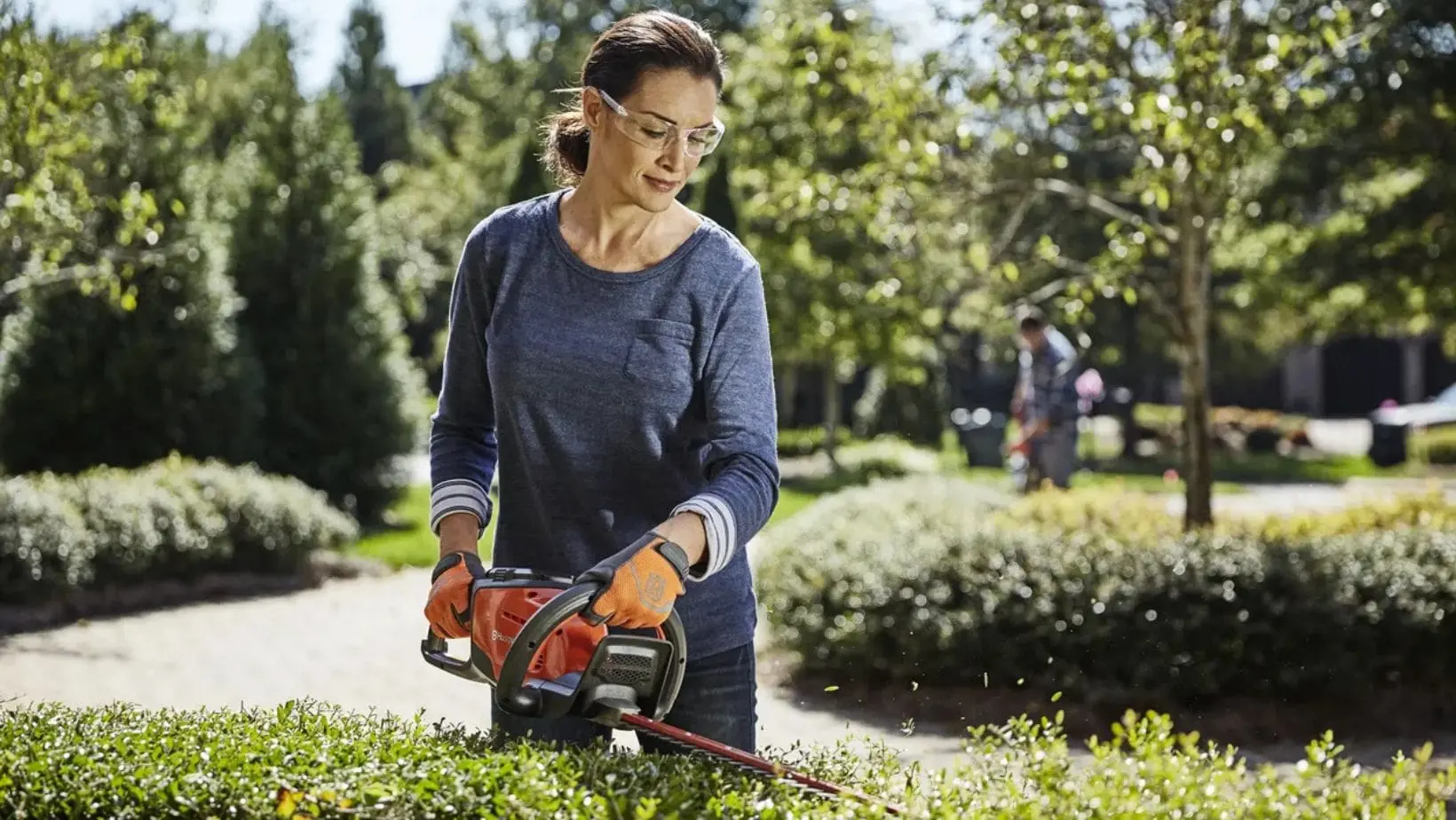 a woman wearing safety glasses, using a hedge trimmer to cut the top of a low-level hedge