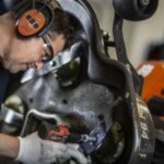 a Husqvarna servicing technician wearing safety glasses and ear defenders, tightening a bolt on the underside of a ride-on mower deck