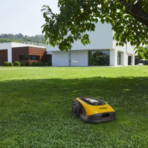 a yellow and grey STIGA robot mower on a lawn outside a large white house, underneath an overhanging branch