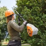 a woman in an orange hardhat, using the STIHL HLA 135 K Cordless Short-shaft Long-reach Hedge Trimmer to trim a tall hedge