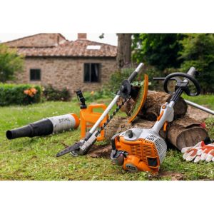 the STIHL KM 56 RC-E Petrol KombiEngine and compatible multi tools resting against 3 logs on a lawn, with a farmhouse in the background