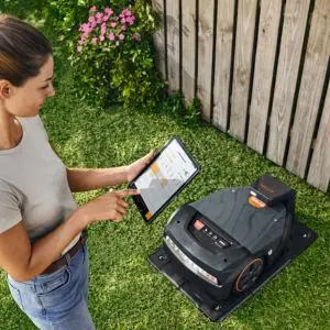 a woman stood next to a black robotic lawn mower, programming it using a tablet