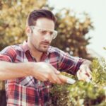 a man wearing a checked shirt and safety glasses, using manual hedge cutters