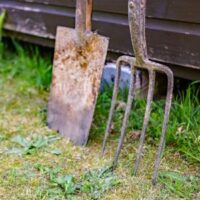 a close up of a rusty garden spade and fork, propped up against a wooden garden building