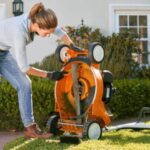 a woman wearing gloves, inspecting the underside of a lawnmower