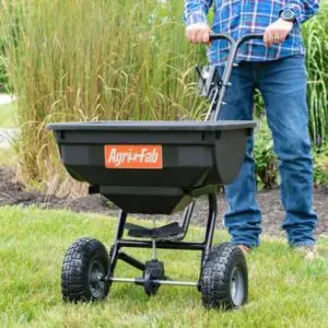 a man stood on a lawn, holding the handle of the AGRI-FAB 45-0532 Pro Push Spreader