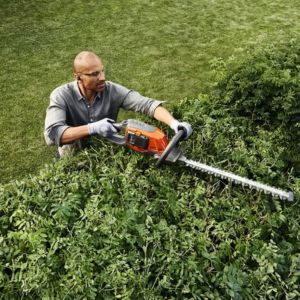 a man wearing gloves and safety glasses, using the Husqvarna 215iHD45 Hedge Trimmer to cut the top of a hedge