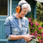 a smiling woman with short grey hair, wearing a denim jacket, ear defenders and safety glasses, using a mobile phone
