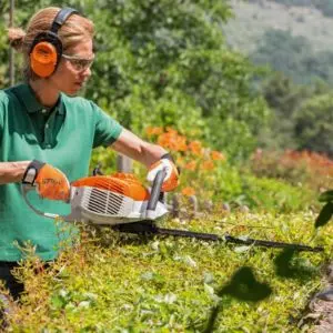 a woman wearing gardening PPE, including ear defenders, safety glasses and gloves, while using a hedge trimmer
