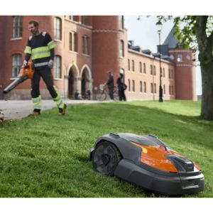 a groundscare worker operating a leaf blower, while a Husqvarna Automower drives down a grassy bank