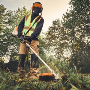 a groundscare worker dressed in PPE, operating a brushcutter in a field, with trees in the background