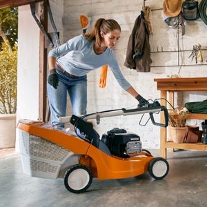 a woman folding the handle of a STIHL lawnmower down into the storage position, inside a well-organised garage with white walls