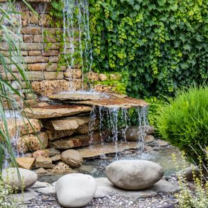 a rustic water feature including large, irregularly shaped stones, surrounded by a hedge, dry stone wall, and shrubs