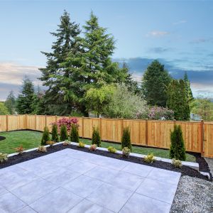 a raised patio in the foreground of a simple garden design consisting of small conifers, a lawn and fencing, with large trees in the background