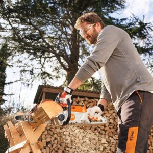 a man using the STIHL MSA 70 Cordless Chainsaw to cut a piece of firewood on a sawhorse, with a full logstore in the background