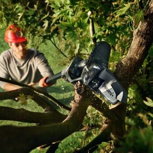 a man stood on a lawn, wearing a red hard hat, using the Husqvarna Aspire™ PE5-P4A Pruning Saw, fitted with an extension pole, to prune high up in a tree