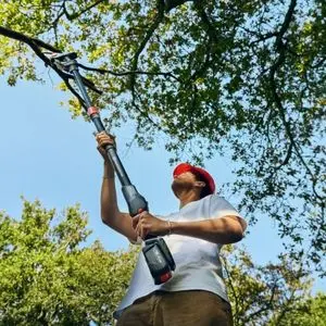 a man wearing a hard hat, using the Husqvarna Aspire™ PE5-P4A Pole Pruner to remove an overhanging branch from a tree