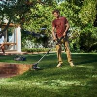 a man wearing a red t-shirt and beige trousers, using the Husqvarna Aspire™ T28-P4A Battery Grass Trimmer to cut the edge of a lawn, with a house and trees in the background