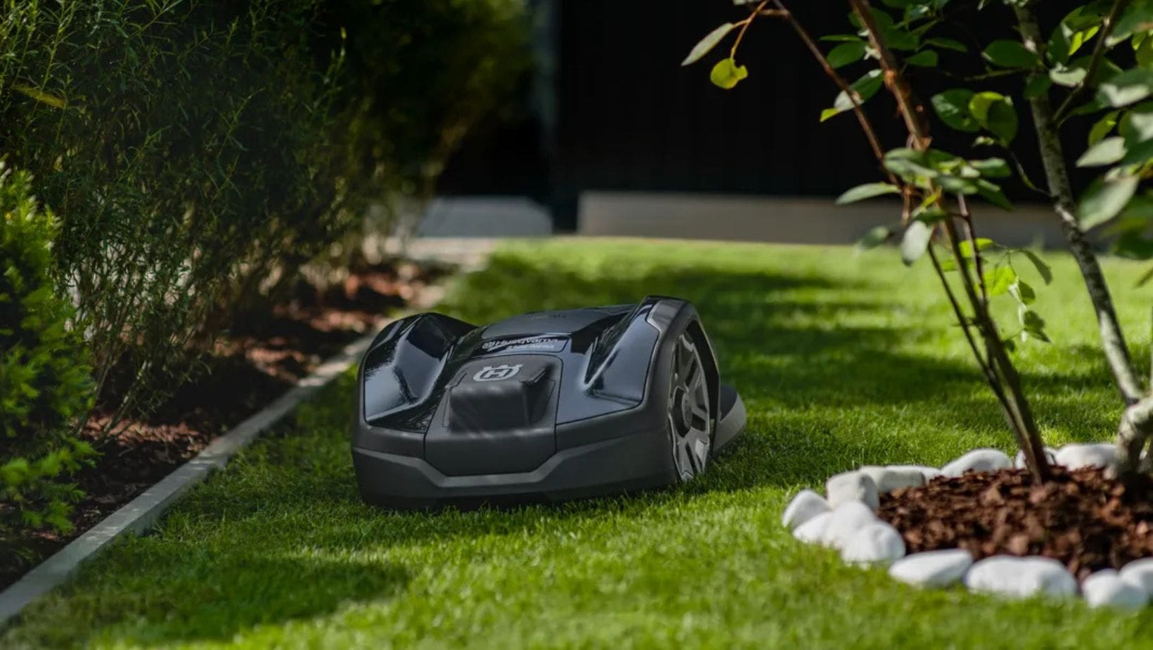 the Husqvarna Automower® 305E NERA Robotic Mower travelling on a lawn, between a border and other plant