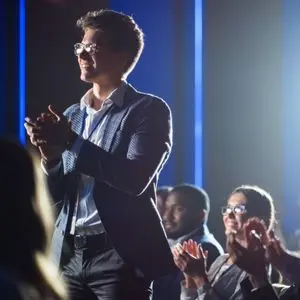 a man wearing a suit and spectacles, surrounded by other smartly dressed people, stood up clapping at an awards ceremony