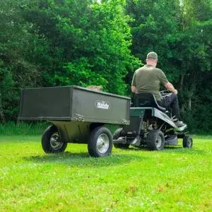 a man using a ride-on mower to tow the Handy THGT500 Towed Steel Tipping Trailer across a lawn, with trees in the background
