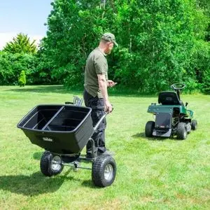 a man in a field, pulling the Handy THTS175 Towed Broadcast Spreader Attachment towards a ride-on mower