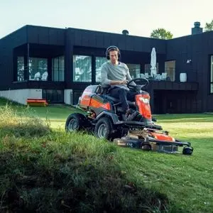 a man driving a Husqvarna out-front ride-on mower across an undulating lawn, with a black, modern building in the background
