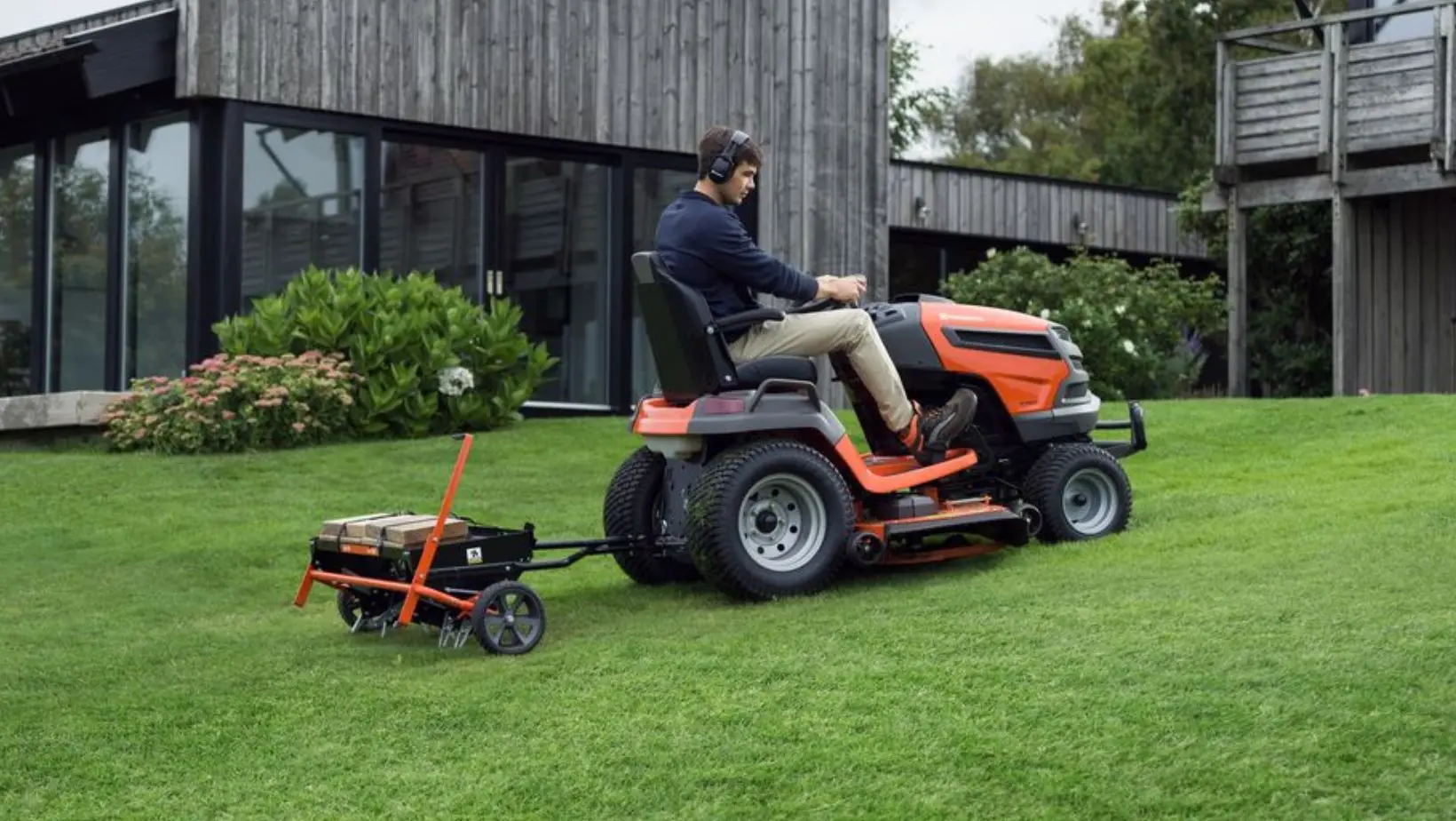 a man using a Husqvarna ride-on mower to tow an aerator attachment up a sloping lawn, with a modern house in the background