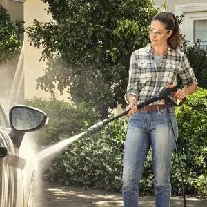 a woman dressed in a checked shirt and jeans, using a pressure cleaner to wash a car