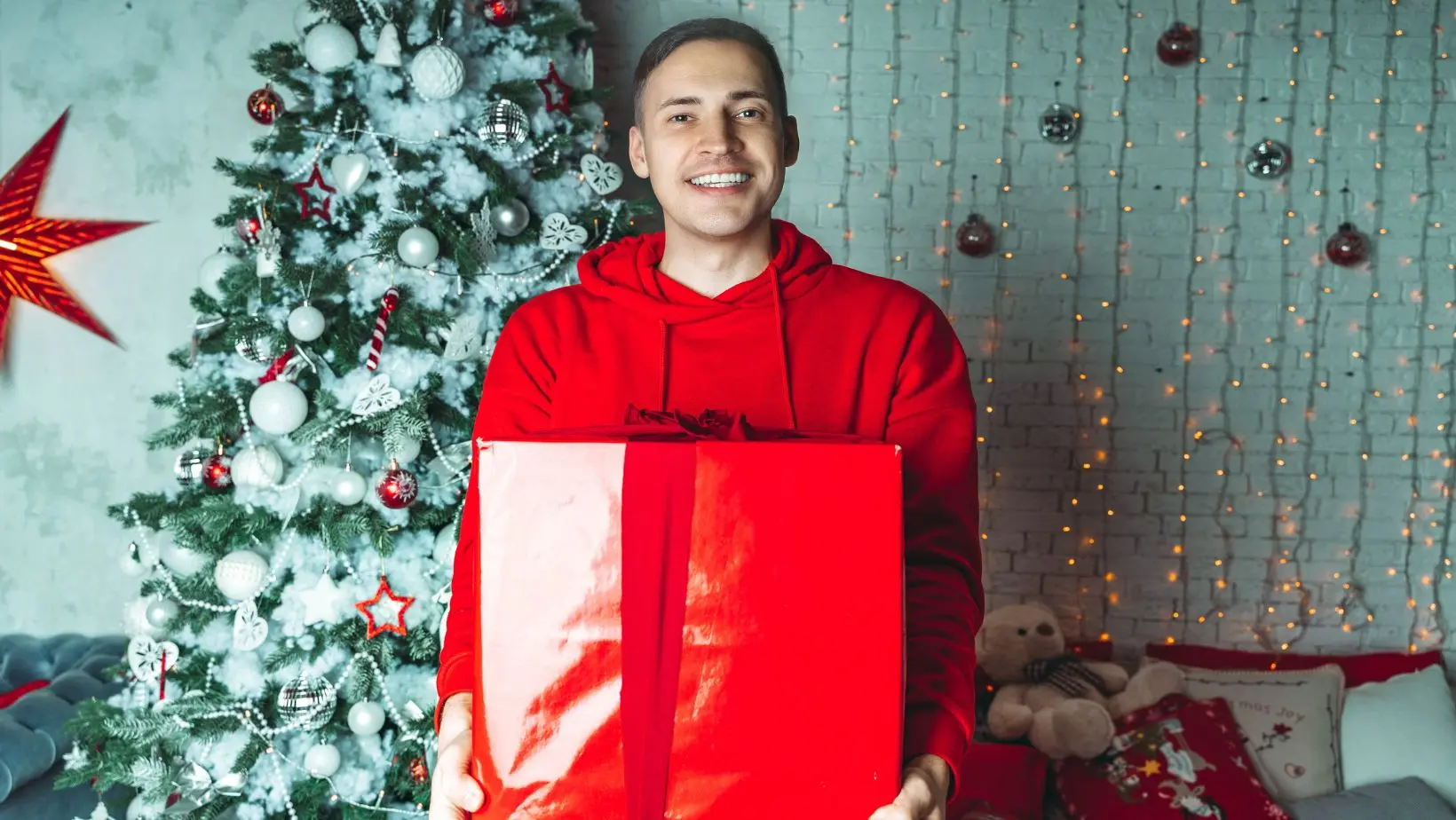 a smiling man dressed in a red jumper, stood next to a Christmas tree, holding a large present in red wrapping paper