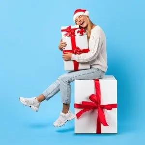 a woman wearing a Santa hat, sat on a large Christmas present that has white wrapping paper and a red bow, holding another similar but smaller present