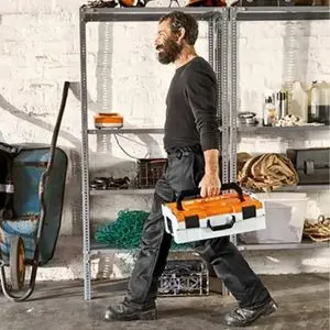 a man with a beard, carrying a battery storage box across a white-walled garage, which is lined with metal shelving