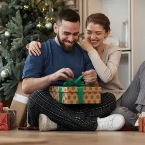 a smiling man and woman, sat cross legged, next to a Christmas tree, as the man opens a Christmas present